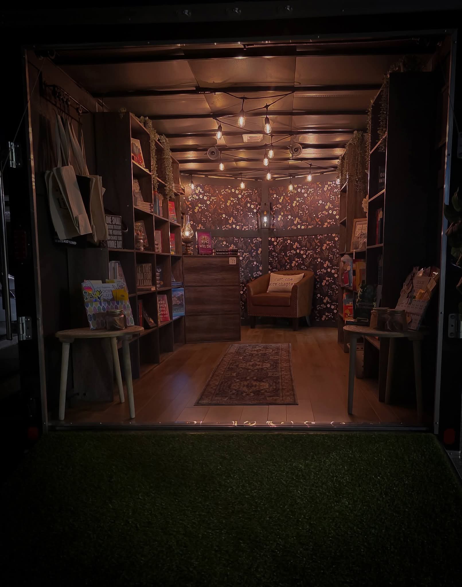 The Ledger & Lamp mobile bookstore interior — warm string lights, curated bookshelves, leather armchair, and dark floral wallpaper inside the traveling trailer