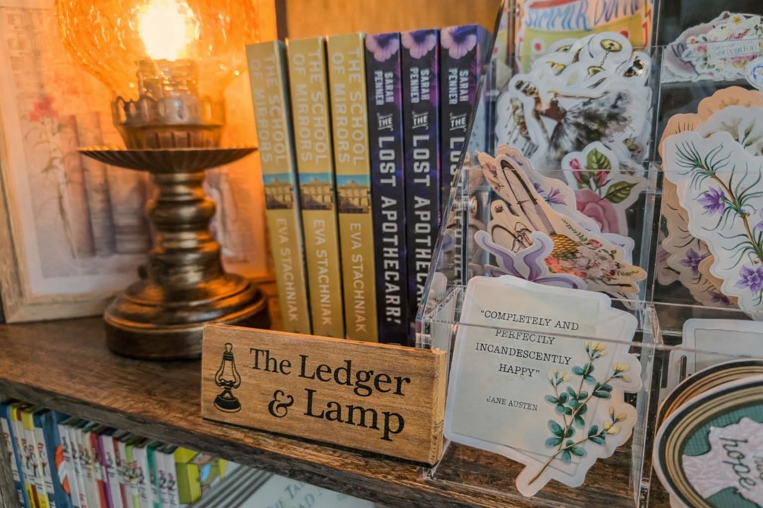 The Ledger & Lamp wooden lantern-logo sign beside literary stickers, including a Jane Austen quote — completely and perfectly incandescently happy