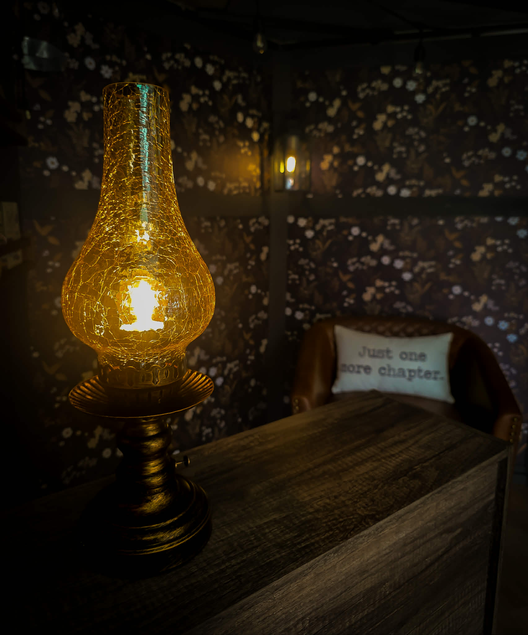 A vintage amber oil lamp glowing softly inside The Ledger & Lamp mobile bookstore, with the Just One More Chapter pillow in the background